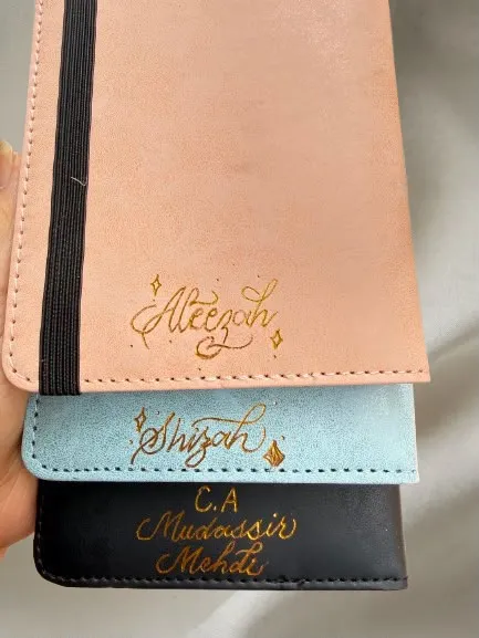 Gold heat-foiled names on pink, blue, and black leather notebooks